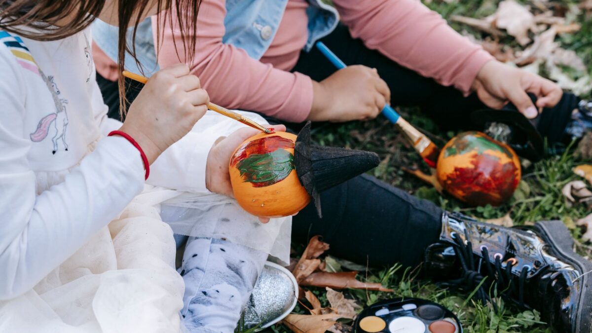 High angle of crop anonymous multiethnic girls sitting on grass and painting pumpkin with aquarelle for Halloween celebration