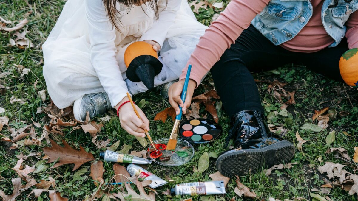 Crop little kids sitting on grass covered with dry foliage and painting on pumpkins for Halloween with brushes