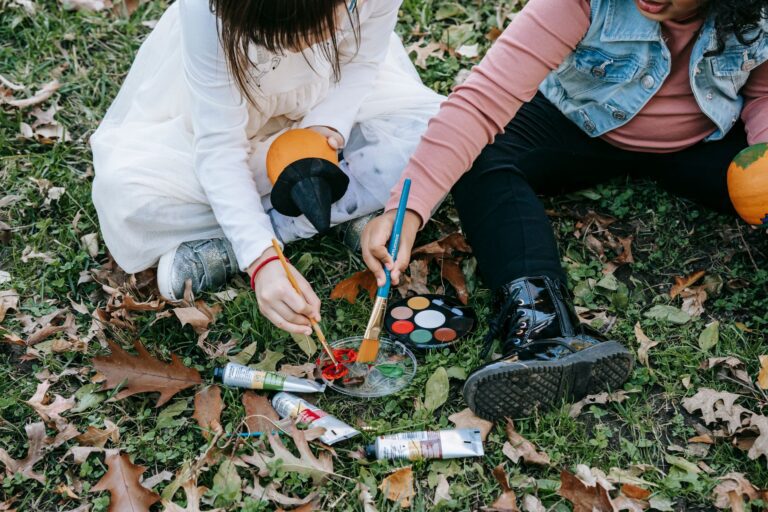 Crop little kids sitting on grass covered with dry foliage and painting on pumpkins for Halloween with brushes