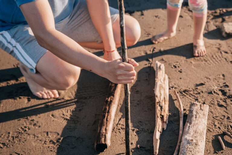 Hands holding a stick, crafting on a sunny beach with driftwood.