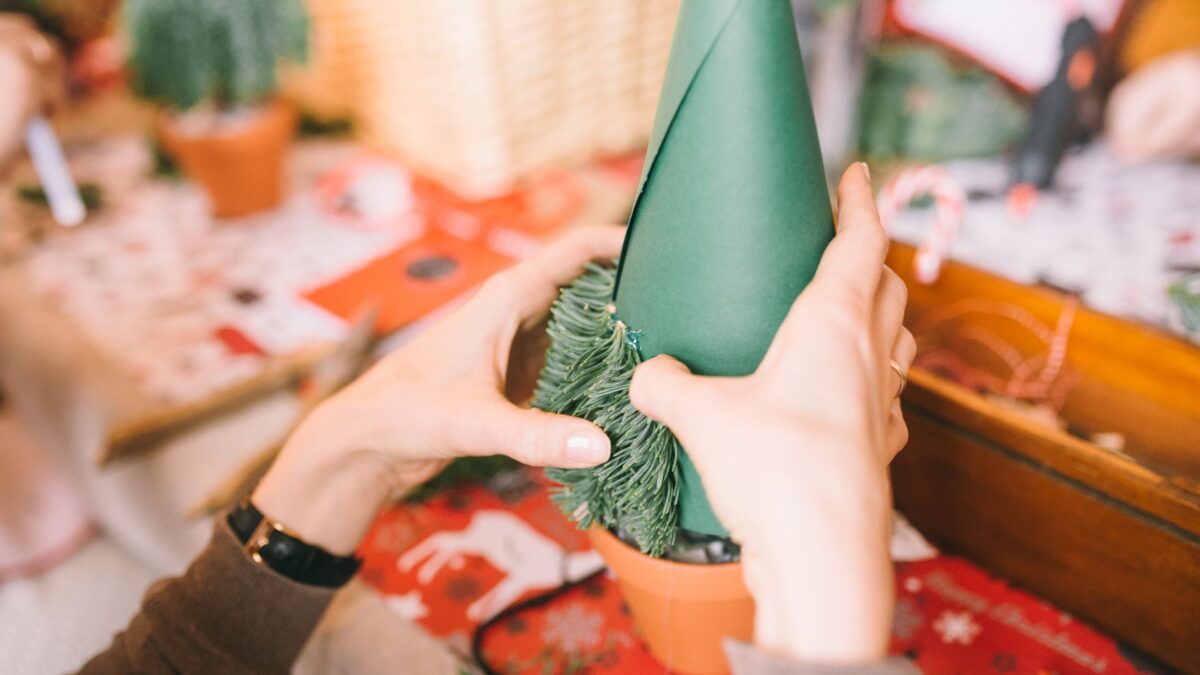 Hands crafting a DIY Christmas tree using green paper and pine leaves for holiday decor.